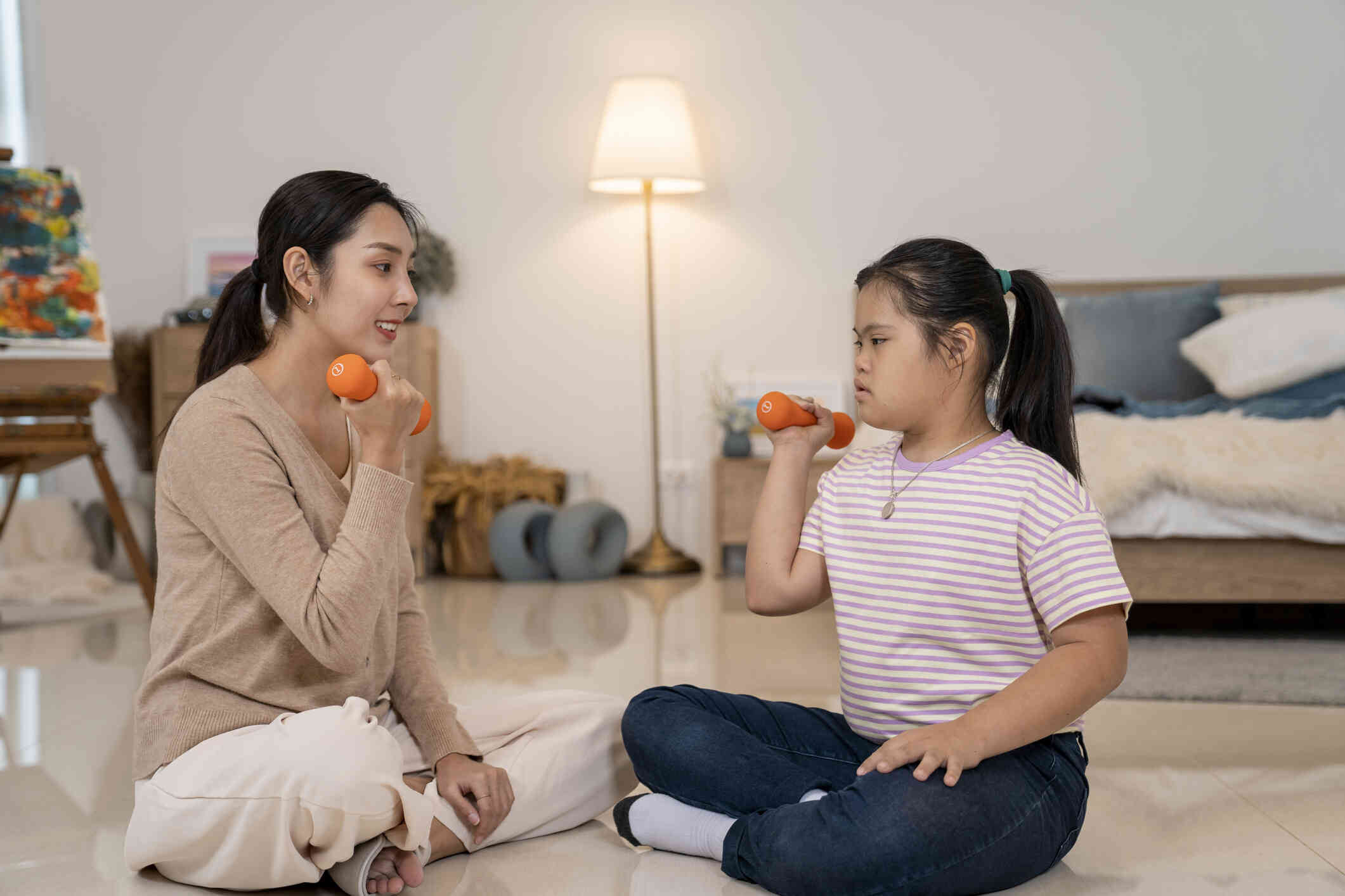 A mother and daughter sit on the floor facing one another while lifting small weights together.